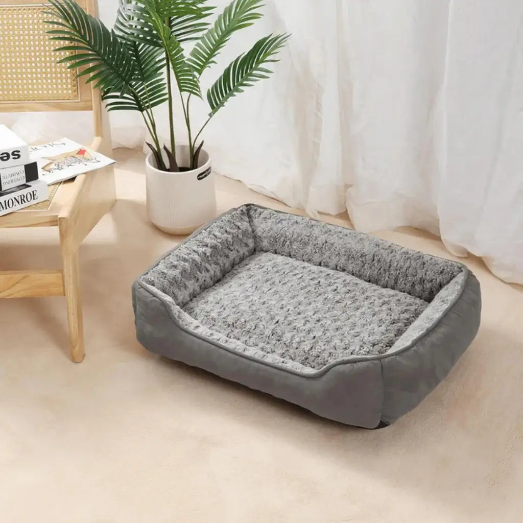 Gray pet bed on a beige floor with a plant and wooden furniture in the background