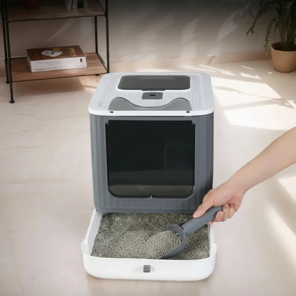 Pet litter box with a scoop on a light-colored floor, with a hand holding the scoop