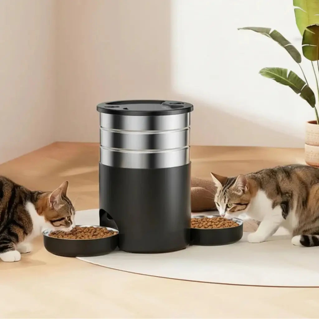 Two cats eating from a black and silver pet food dispenser on a wooden floor